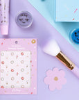 Makeup products for kids including a brush, nail stickers, lipgloss and face paint and eye shadows on a pastel blue and purple background
