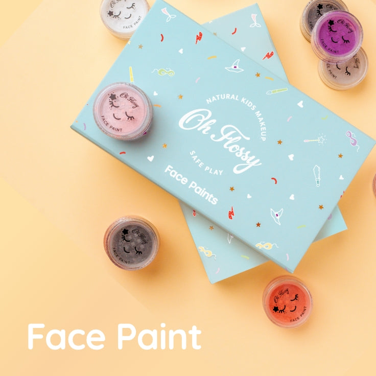 A flat-lay of a light blue face paint palette box labeled 'Oh Flossy Safe Play Face Paints' with several small round pots of face paint arranged around it on a pale yellow background. The text 'Face Paint' appears at the bottom left.