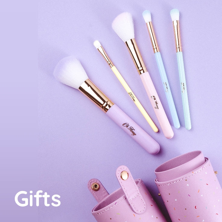 A set of pastel makeup brushes with white bristles and rose gold ferrules, arranged on a lavender background next to a matching pink cylindrical case. The brushes have pastel purple, pale yellow, light pink, and light blue handles. The text in the bottom left corner reads 'Gifts'