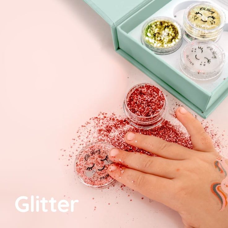 A kid's hand is shown touching red glitter spilled on a light pink surface, with small clear pots of glitter and a mint box in the background. The word 'Glitter' appears in white text at the bottom left.