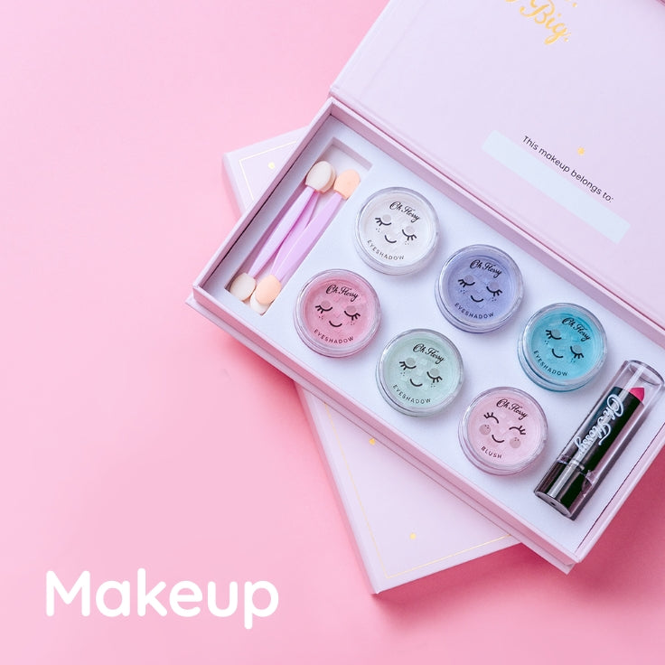 A pink makeup gift set displayed in an open box on a pink background. The set includes six small round eyeshadow pots with cute face drawings, two pink dual-ended applicators, and a pink lipstick tube. The text in the bottom left corner reads 'Makeup'