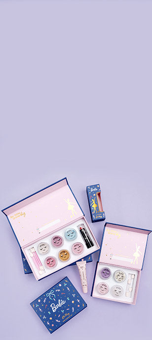 A Barbie-themed kids make-up gift set arranged on a light lavender background, including open pink and blue boxes with eyeshadows, glitter, blush, lipstick, lip gloss and prime.