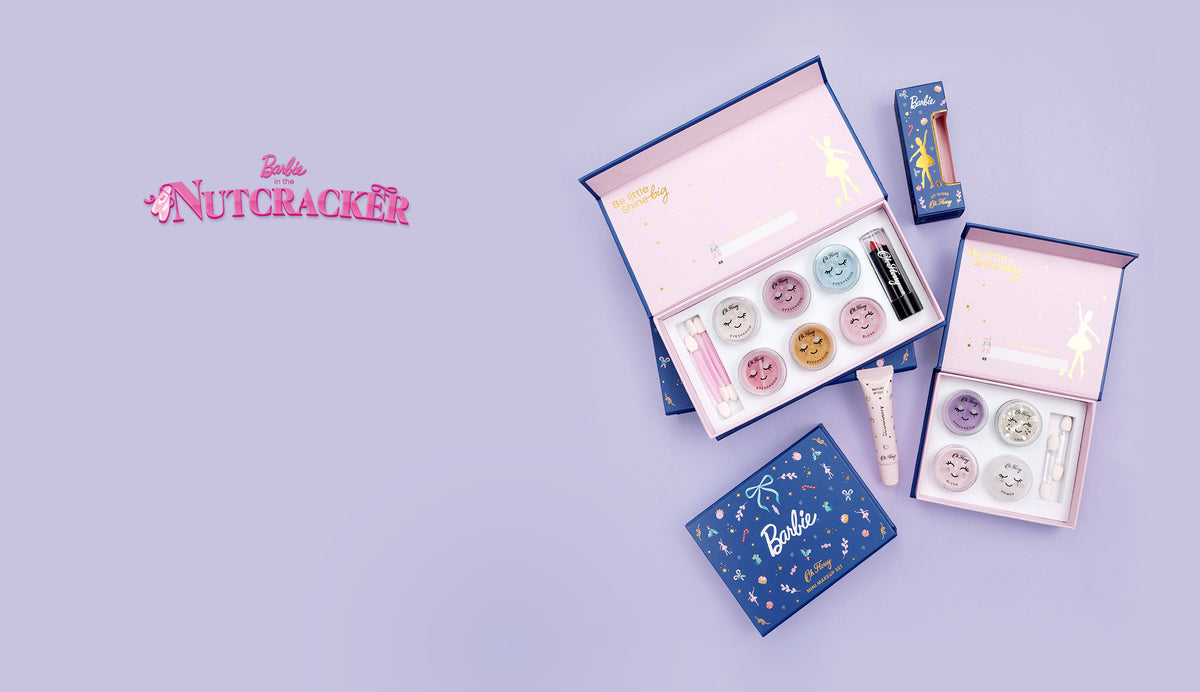 A Barbie-themed kids make-up gift set arranged on a light lavender background, including open pink and blue boxes with eyeshadows, glitter, blush, lipstick, lip gloss and prime.