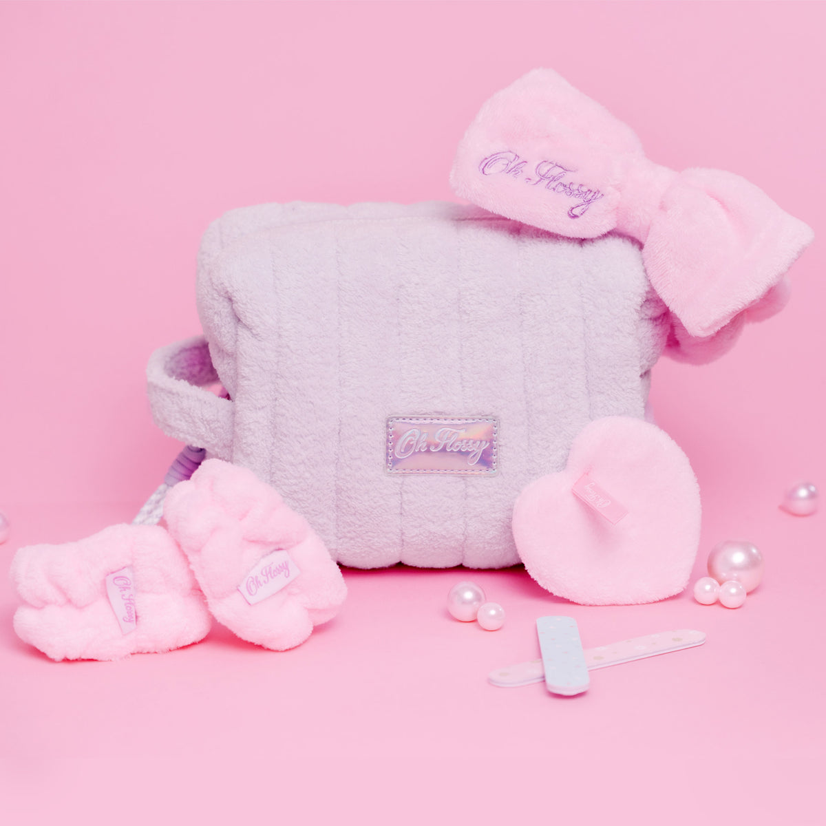 A pink and lavender gift set featuring a quilted lilac cosmetic bag, a matching bow headband, wrist cuffs, a heart-shaped makeup remover pad, and nail files, all arranged on a solid pink background. The brand name 'Oh Flossy' is embroidered on the bow and label.