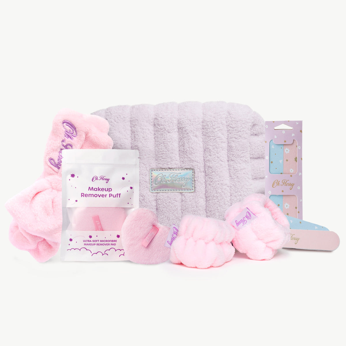 A pink and lavender self-care gift set on a white background, including a lavender plush bag, a makeup remover puff, a pink cosmetic headband and matching wrist cuffs and a nail files.