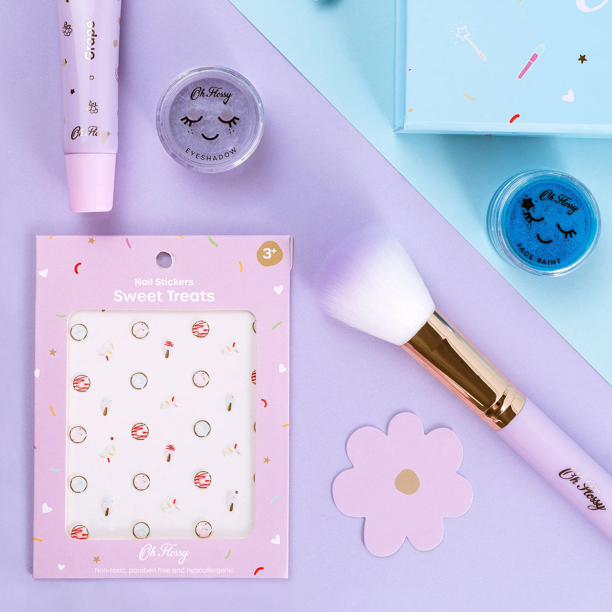 Makeup products for kids including a brush, nail stickers, lipgloss and face paint and eye shadows on a pastel blue and purple background