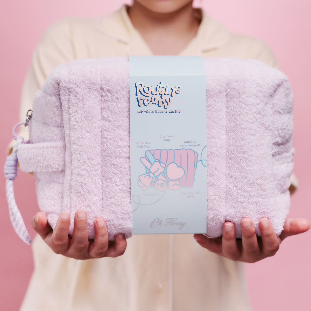 A girl in lemon pajamas holds a light purple plush cosmetic bag with a matching wrist strap. The pouch has a central paper label that reads 'Routine Ready' and 'Cosmetic Essentials Kit' with a cute illustration.