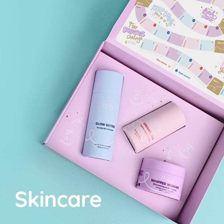 A pastel skincare gift set arranged inside an open pink box on a light aqua background. The set includes a tall light blue bottle labeled Glow Getter Cleanser, a pale pink labelled Dreamy Drew Moisturiser, and a lavender jar labeled Whipped Wonder body customer, with a printed insert  7-day skincare challenge visible inside the box.