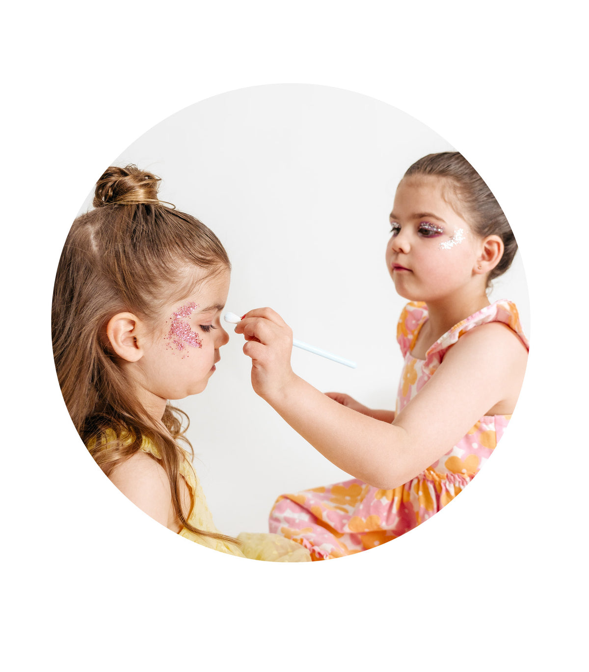 Two young girls playing makeup, one applying pink glitter to the other's forehead with a small Oh Flossy brush.